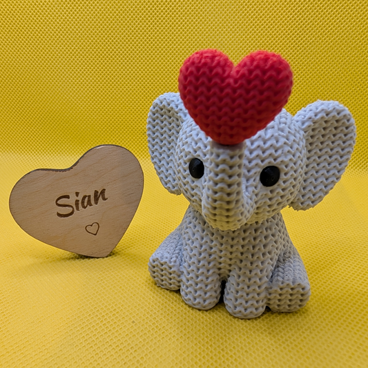 Grey knitted-style elephant figurine with red heart beside a personalised wooden heart engraved Sian